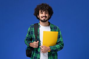 front-view-male-student-green-checkered-shirt-wearing-black-backpack-holding-files-blue-wall-scaled